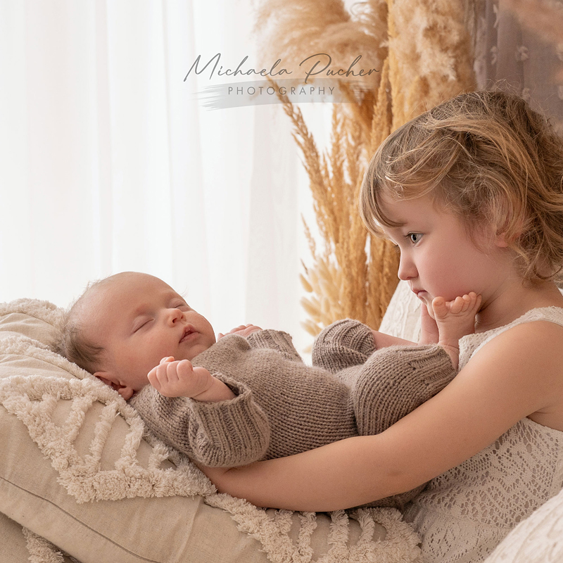 Ein kleines Mädchen hält ihr schlafendes Neugeborenes vorsichtig im Arm, beide liegen auf weichen, beigen Kissen; warmes Tageslicht fällt durch den Vorhang.